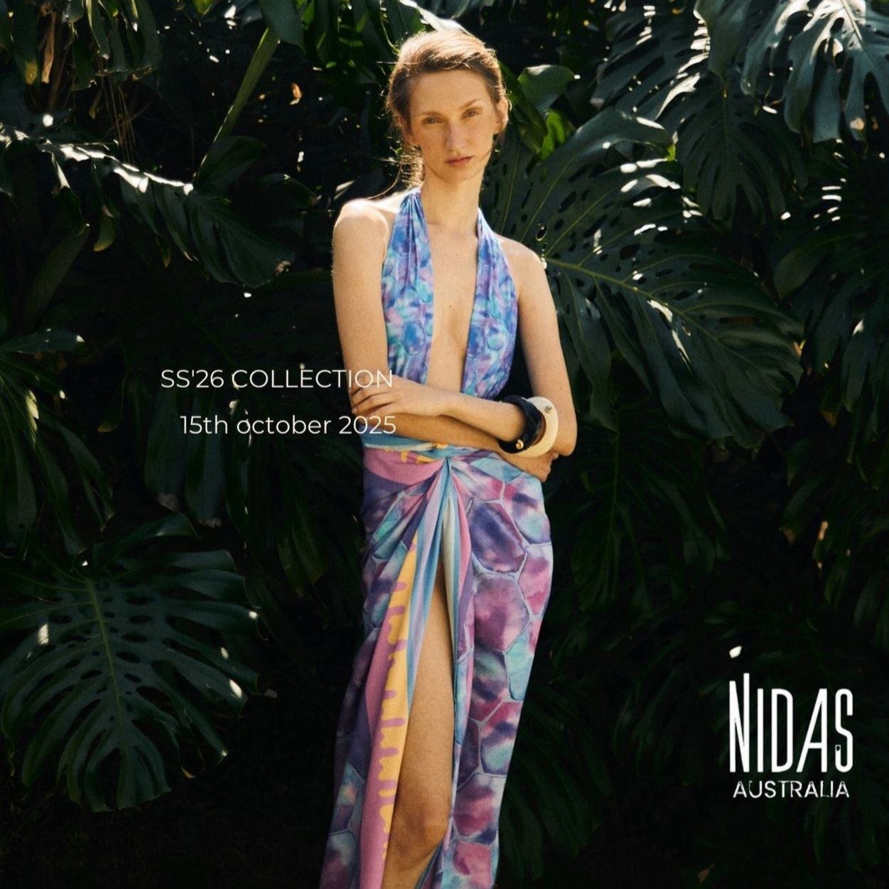 Woman in a colorful one piece and sarong
 standing among large green leaves with 'Nidas Australia' branding.